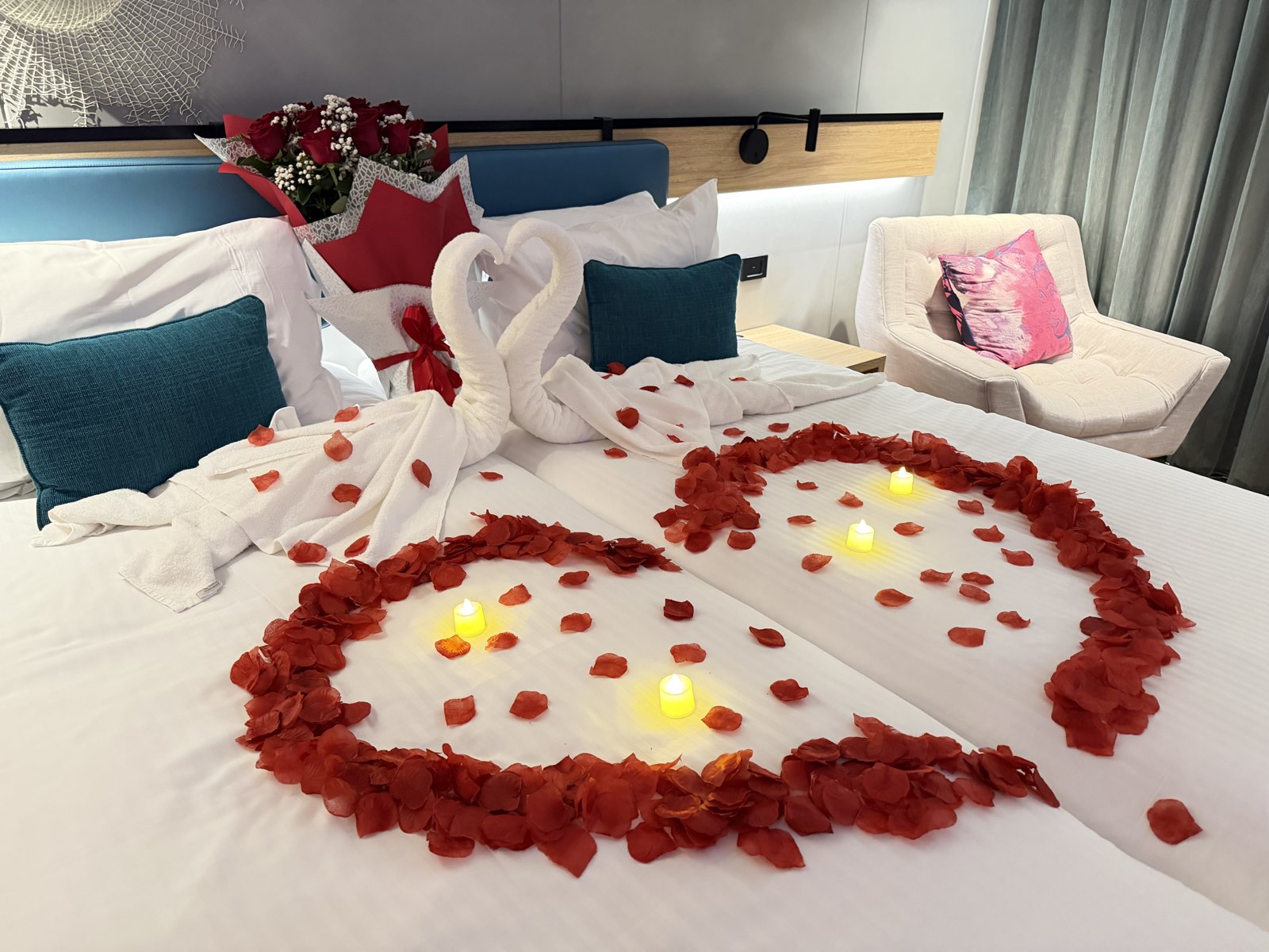 斜めから見た客室のベッドと、バラの花びらのハート、タオルのスワン、枕元の花束
