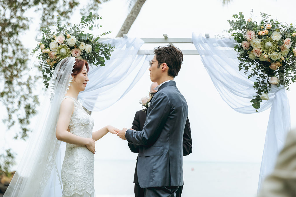 花と白い布で飾られたアーチの前で向き合う新郎新婦