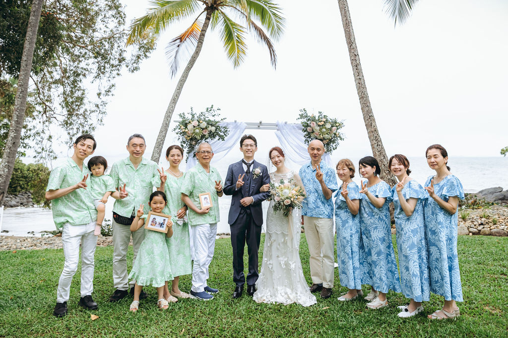 ヤシの木の下で水色の衣装のご家族と並ぶ新郎新婦の集合写真